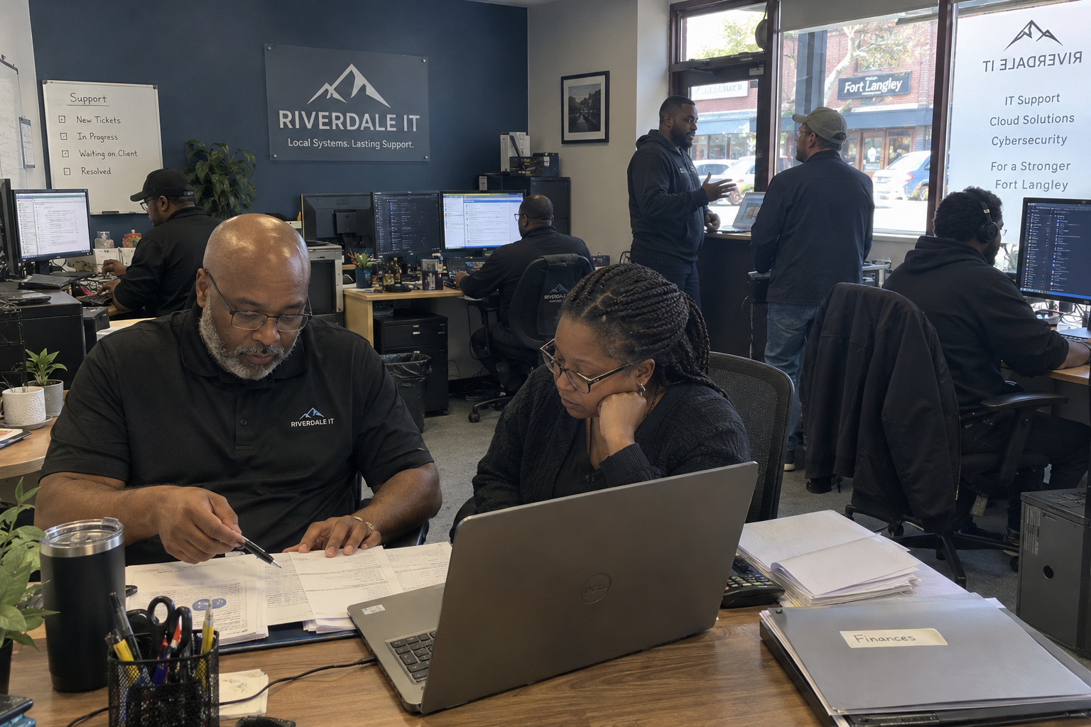 MSP owner dependency discussion with financial papers and a laptop while technicians work at nearby desks.