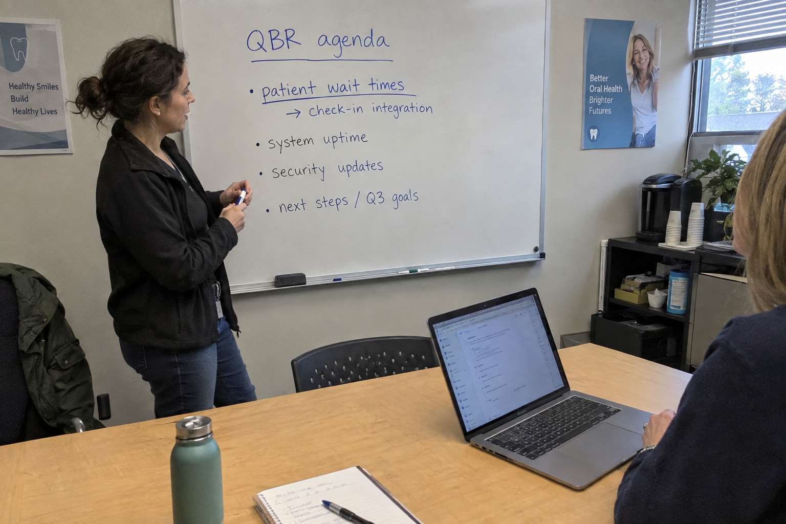 A woman on a phone call points at a tablet while a technician working on an open laptop looks over at it.