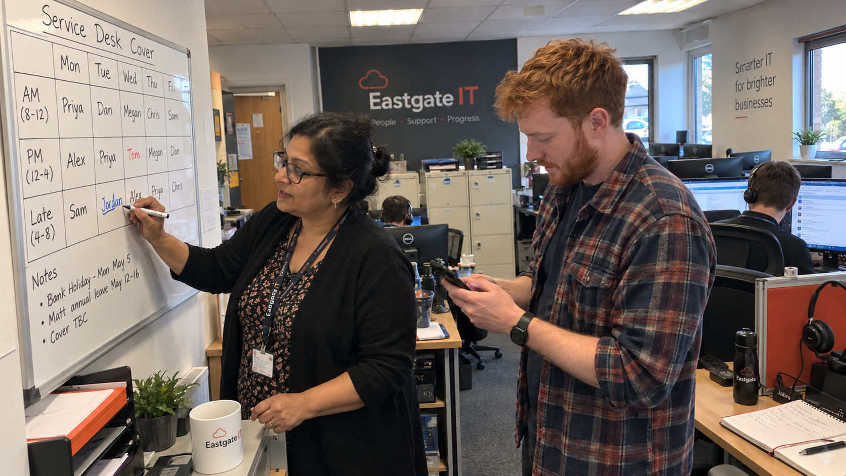 A service lead updates the shift-coverage whiteboard while a technician checks a phone beside her.