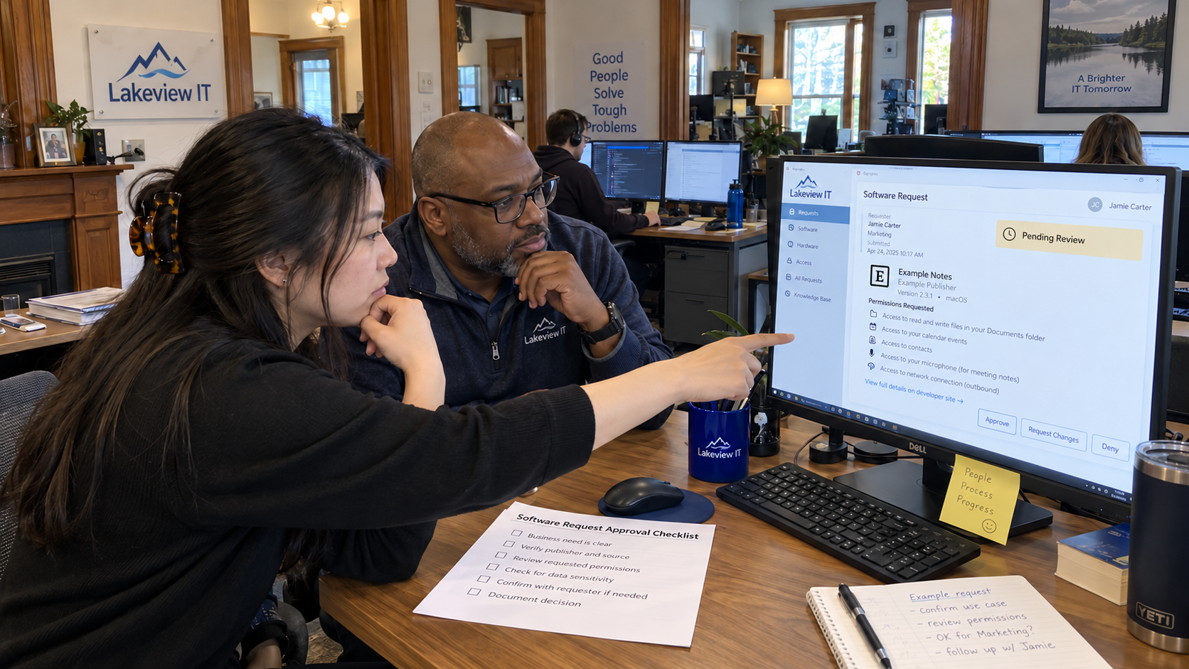 Two colleagues review app permissions on a monitor beside a software approval process checklist.