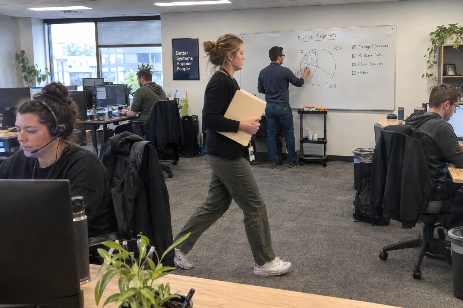 A man in a blazer carries a revenue report across an open office while a technician works at a laptop.