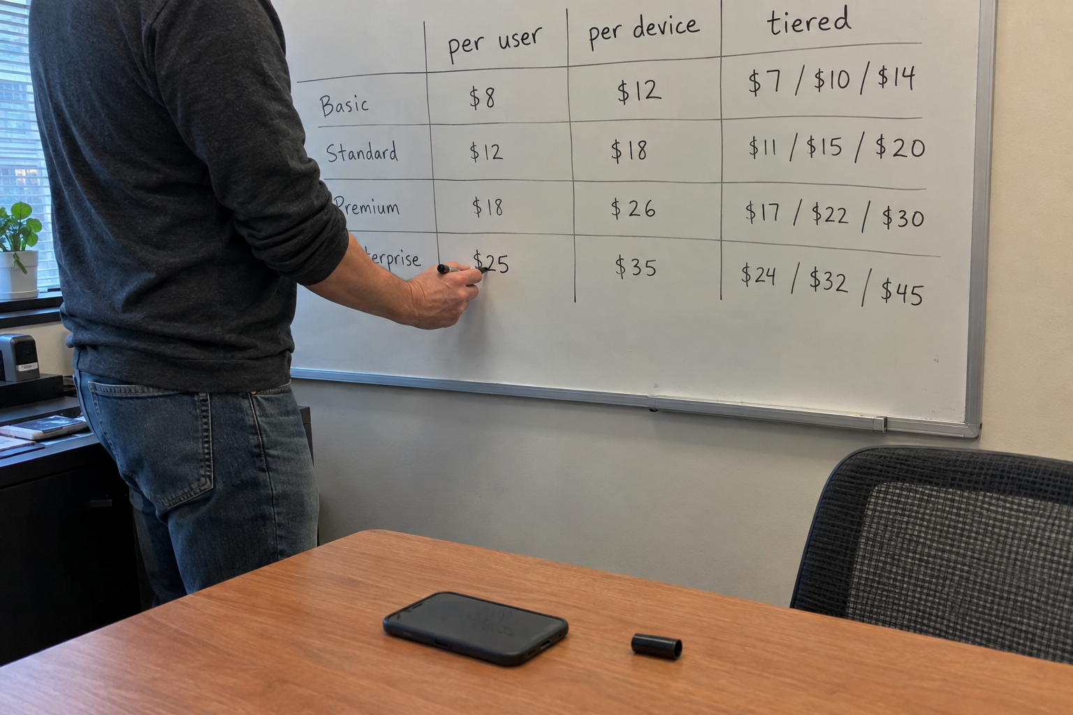A man points a pen at columns of figures on a monitor while a colleague on a headset works at the next desk.